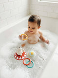 A baby sitting in a bubble-filled bathtub playing with colorful bath toys, including a yellow ring, a red and white shark toy, and a blue star toy.