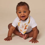 A smiling baby sitting on a beige studio floor wearing a white short-sleeve bodysuit and a white bib with a warm-toned animal and star print.