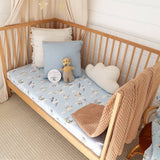 Wooden crib with baby bedding, including a blue crib sheet, white pillow, teddy bear, and brown blanket.