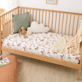 A wooden baby daybed with a fitted sheet printed with a playful fox and rabbit pattern, styled with a green pillow, a cloud-shaped cushion, a plush toy, and a cream throw blanket in a bright nursery setting.