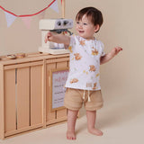 A toddler stands barefoot in front of a toy kitchen setup, wearing a white short-sleeve top with a teddy bear print and tan shorts. The scene includes a toy counter, small appliance, and pastel bunting against a warm beige background.