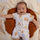 A baby lying on a brown quilted crescent pillow, wearing a white zip-up baby romper printed with cute cartoon sheep and yellow accents. The background consists of cozy cream-colored knitted blankets.