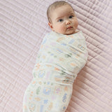 A baby swaddled in a light cream muslin blanket with a pastel multicolor sea life pattern, lying on a pink quilted mattress.