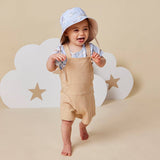A baby wearing a light blue sun hat with a star pattern, a light blue striped shirt, and beige overall overalls, standing barefoot in front of a beige background with white cloud cutouts.