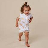 A toddler girl wearing a white short-sleeve zip-up romper with a yellow and brown animal print, walking barefoot against a solid beige background.