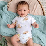 A baby lying on a mint green blanket, wearing a white short-sleeve bodysuit with a print of cartoon bunnies and chicks. The baby is sitting in a woven bassinet with a beige cushion.