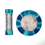 A teal and blue marbled shaker candle shown in two views: a tall cylindrical tube with a glitter-filled center and a round plate-like container with a clear top containing small star and flower confetti, on a white background.