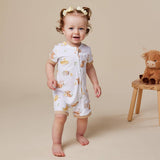 A baby girl wearing a white short-sleeve footless romper with a yellow and brown animal print, standing barefoot on a beige studio floor next to a small wooden stool with a plush toy.