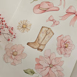 A flat-lay view of a watercolor-style print or card featuring a tan cowboy boot, pink flowers, daisies, bows, and a cowboy hat on a warm off-white background, with small dried pink flowers placed along the left edge.
