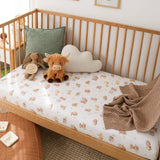 A wooden crib or daybed styled in a nursery with a white fitted sheet printed with a small teddy bear pattern, a brown knit throw blanket, a green pillow, and plush toys including a cow.