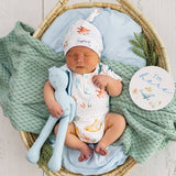 A newborn baby is lying in a woven basket bassinet, wearing a white one-piece bodysuit and matching hat with orange sea-creature prints. The baby is holding a light blue plush bunny toy.