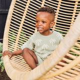 Child sitting in a wicker chair wearing a green sweater with palm tree patterns.