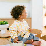 Child wearing a white shirt with yellow patterns sitting on a wooden floor.