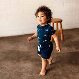 A toddler walking barefoot indoors wearing a navy blue short-sleeve romper and matching shorts with a white space-themed print of planets and stars.
