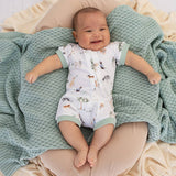 A baby lying on a textured mint green blanket, wearing a white footless onesie with a woodland animal print and sage green trim.