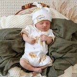 A sleeping newborn baby wearing a white zip-front bodysuit and matching beanie hat with a teddy bear print, nestled in a dark olive green blanket on a cozy textured blanket background.