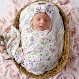 A sleeping newborn baby wrapped in a white muslin swaddle blanket with a colorful floral print, wearing a matching bow headband, resting inside a woven wicker basket on a pink blanket.