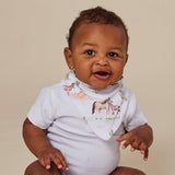 A smiling baby wearing a white ribbed short-sleeve bodysuit and a white bib with a horse and donkey animal print, posed against a solid beige background.