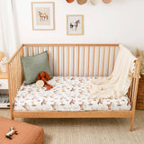 A light natural wood crib with a fitted sheet printed with a whimsical horse and cow pattern, styled with a green pillow, a plush toy, and a cream throw blanket in a bright nursery setting.