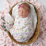 A sleeping newborn baby swaddled in a white floral blanket with a pink bow headband, resting inside a woven basket on a pink blanket.