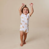 A toddler girl wearing a white sleeveless sleep sack with a cute teddy bear print, standing barefoot against a solid beige background with her arms raised in the air.
