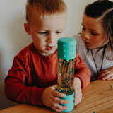 Two children looking at a turquoise sensory bottle with glitter on a wooden table.