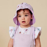 A young child wearing a light purple floral sun hat with drawstrings and a matching lavender overall dress, posed against a solid beige background.
