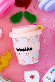 White cup with peach designs and a pink lid on a pink background with sprinkles and toys.