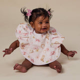 A baby girl sitting on a beige studio floor wearing a white bib with pink bows and cowboy boot prints, with her hair in two pigtails.