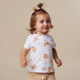 A toddler girl with a small hair bun wearing a white short-sleeve henley shirt with a teddy bear print and khaki pants, standing against a solid beige background.