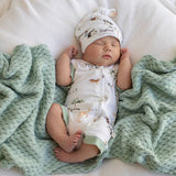 A sleeping newborn baby wearing a white zip-front bodysuit and matching beanie hat with small animal prints, lying on a white textured blanket between two mint green chunky knit throws. The hat shows the brand name 'Simply Happy'.