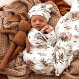 Newborn baby wrapped in a floral blanket with a brown stuffed toy, lying on a soft surface.