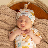 A sleeping newborn baby wearing a matching cream-colored onesie and knotted hat set with a colorful food-themed print, lying on a textured beige blanket inside a woven basket.
