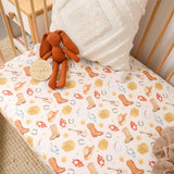 A fitted crib sheet with a western-themed print featuring cowboy boots, horseshoes, lasso rings, and stars on a cream background, styled in a nursery with a plush bunny toy, white textured pillow, and wooden crib.