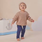 Child wearing a beige sweater and blue pants standing in front of a white and gray toy boat.