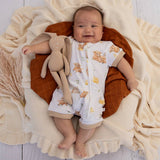 A baby lying on a cream knit blanket with a brown pillow, wearing a white short-sleeve footless romper printed with teddy bear illustrations. The baby is holding a beige plush bunny toy.