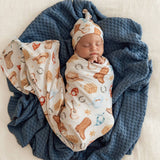 A newborn baby is swaddled in a cream blanket with a western boot and horseshoe print, wearing a matching hat, lying on a textured blue knit blanket.