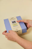 A person's hands holding a light blue and white product box labeled 'BIBS Baby on the Way' against a solid cream-colored background.