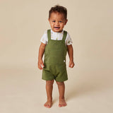 A smiling toddler standing barefoot against a warm beige studio background, wearing olive green overall-style shorts over a white short-sleeve shirt with a small animal print.