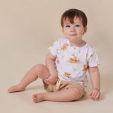 A baby sitting on a beige studio floor wearing a white short-sleeve t-shirt with a brown bear print and tan shorts.