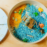 Sensory bin with blue and yellow sand, toys, and a magnifying glass on a wooden surface.