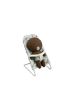 A small brown teddy bear wearing a light green striped diaper and pacifier, seated in a white baby lounge chair with a white star-patterned fabric, isolated on a solid black background.