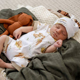 A sleeping newborn baby wearing a white zip-front onesie with a woodland animal print and a matching white beanie hat, lying on a dark green blanket next to a brown plush toy.