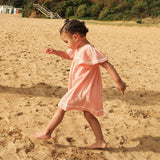 A young child walking barefoot on a sandy beach, wearing a light pink short-sleeve dress with white ruffled trim.