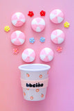 A pink paper coffee cup with a peach pattern and a pink band reading 'bbcióno' in black text, arranged on a pink background with pink-and-white round decorations and small flower embellishments.