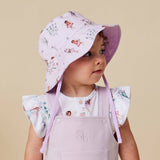 A young child wearing a light lavender sun hat with a whimsical fairy and floral print, paired with matching lavender overalls and a white top with a matching print, set against a solid beige background.