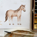A watercolor-style drawing of a light brown horse on white paper, shown with a paintbrush and a watercolor palette in a studio setting.