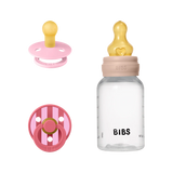 BIBS baby bottle and pacifier set on a white background, featuring a clear bottle with a beige collar and yellow nipple, alongside a pink pacifier and a pink-and-white striped pacifier.