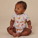 A baby sitting cross-legged wearing a white short-sleeve bodysuit with a cowboy-themed print, photographed against a solid beige studio background.
