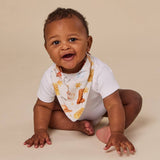 A baby wearing a white short-sleeve bodysuit and a white patterned bib with yellow stars and orange-brown animal illustrations, sitting on a plain beige studio background.